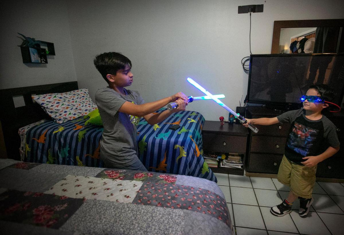 Juan David Acuña, de 13 años, lucha contra su hermano menor, Mateo, de seis años, con unos coloridos sables de luz junto a la "cama de dinosaurio" que Mateo recibió de los donantes del Wish Book en las pasadas fiestas. Mateo compartía la cama con sus padres antes de la donación de la nueva cama, que venía con sábanas, colcha y almohada de dinosaurio a juego, lo que le encantó.
