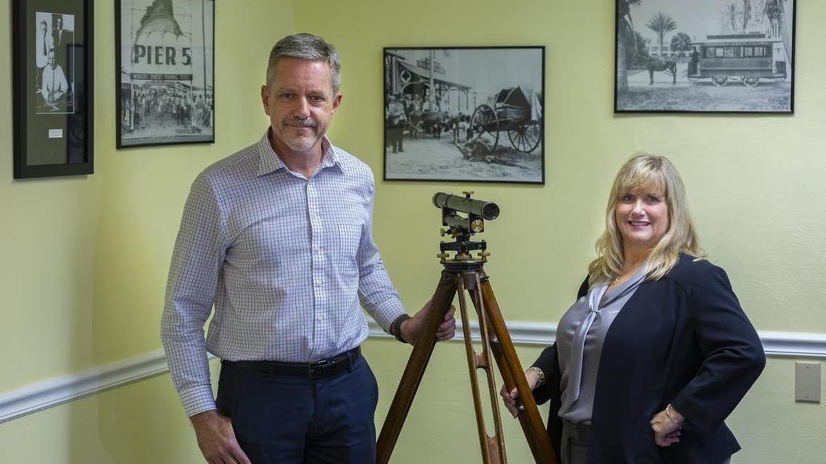 Mike Bartholomew, president of Biscayne Engineering, left, and Melissa Bolton Calabrese, CEO of Biscayne Engineering, in their offices near the Miami River, with an antique surveyor’s tripod. On the walls are images of South Florida in the 20th century.