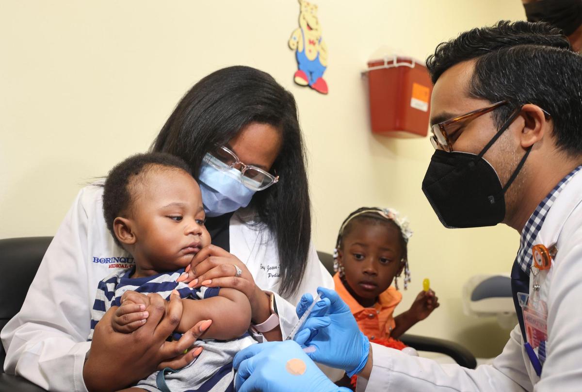 El Dr. Nizar Dowla, a la derecha, administra una vacuna contra COVID-19 a Deji Adebayo, de nueve meses, mientras su madre, la Dra. Emy Jean-Marie, y su hermana, Emiola Adebayo, de tres años, observan. Jean-Marie es médico en el Centro de Atención Médica Borinquen de Miami, donde la secretaria adjunta de salud del Departamento de Salud y Servicios Humanos de Estados Unidos, Rachel Levine, y la alcaldesa de Miami-Dade, Daniella Levine Cava, asistieron a la campaña de vacunación el martes 28 de junio de 2022.