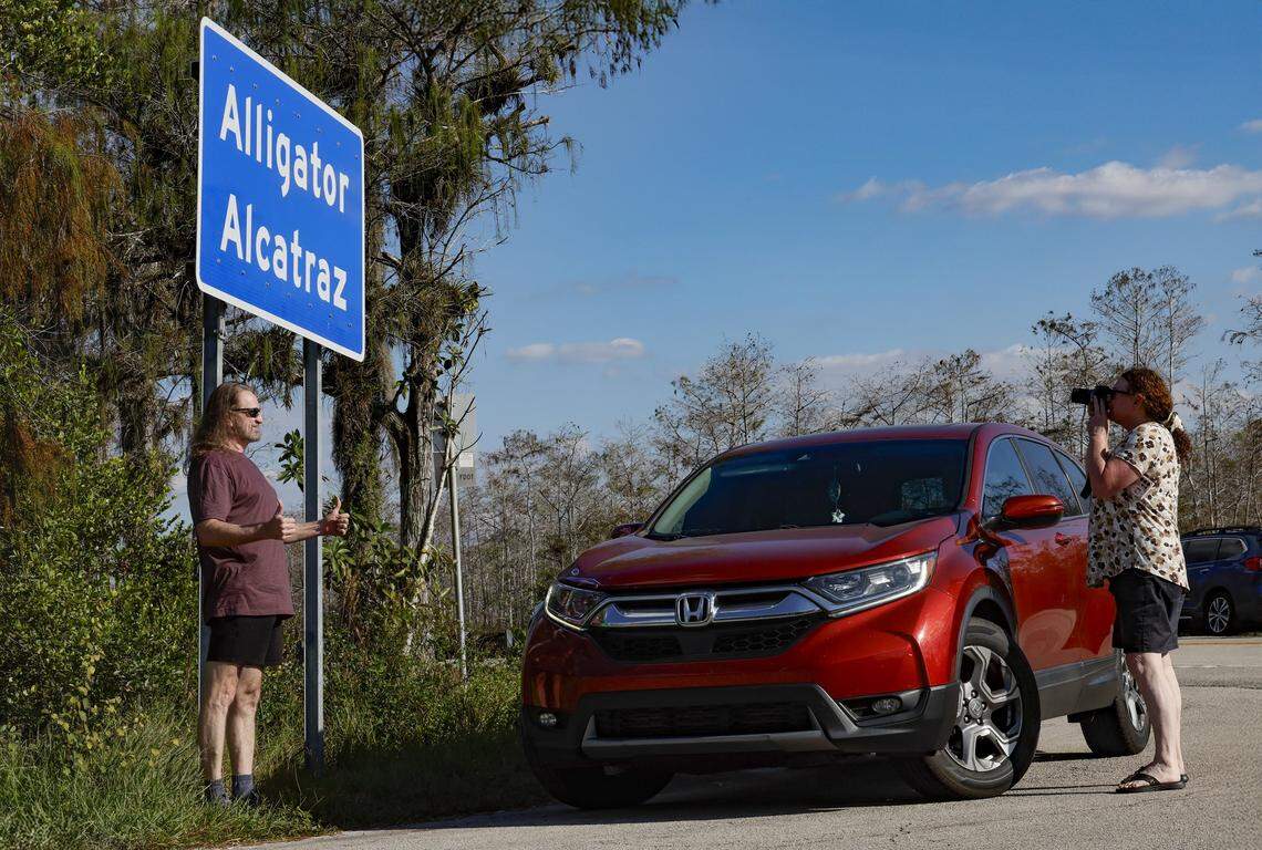 Flashing a thumbs up and posing for photographs is a man standing underneath the sign at the front entrance to Alligator Alcatraz, an immigration detention facility along Tamiami Trail, and located at Dade-Collier Training and Transition Airport inside Big Cypress National Preserve in Ochopee, Florida on Wednesday, November 19, 2025.  