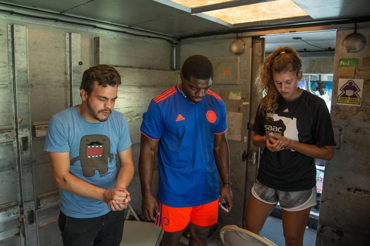 Christian Delgado, left, says a prayer with University of Miami student athletes Robert Burns, center, and Hannah Marwede, inside the Second Spoon food truck before giving out food in  Overtown on Saturday, July 7, 2018.