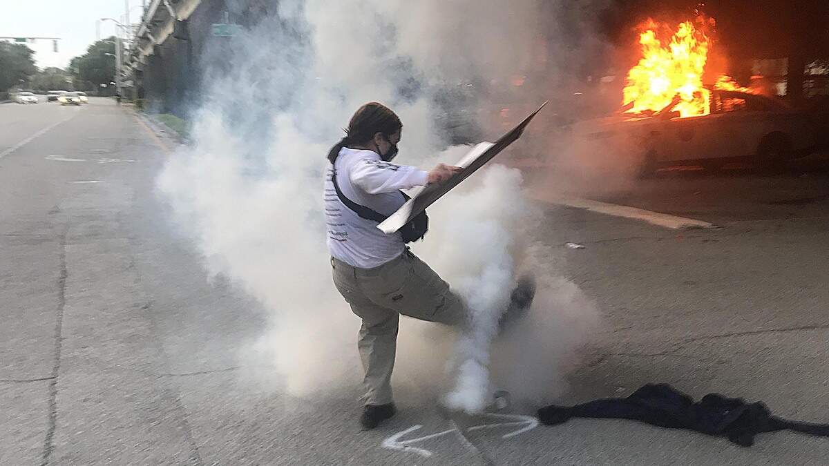 Un auto patrullero de Miami arde en el fondo mientras un manifestante trata de patear una granada de gas lacrimógeno junto a la sede de la Policía de Miami, el sábado 30 de mayo de 2020.