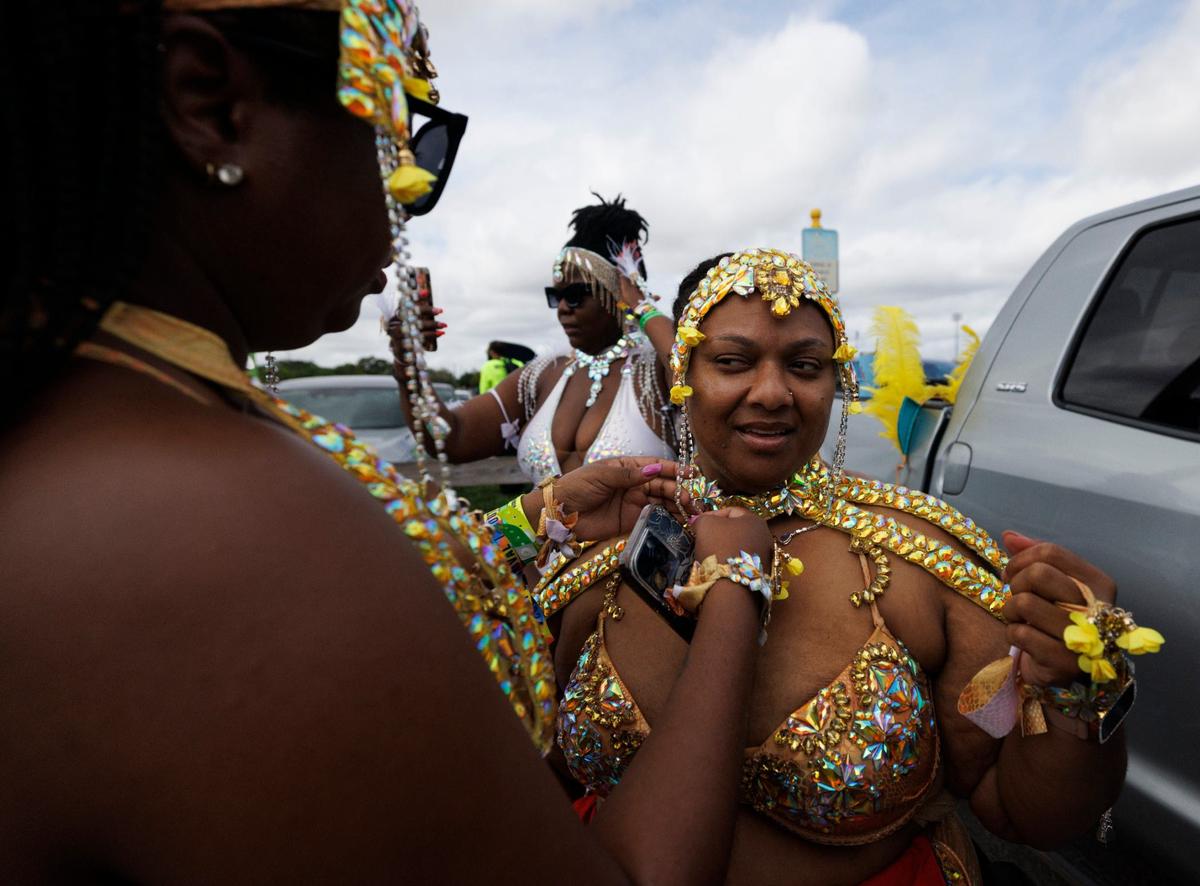 Nell, left, ties Marjorie’s costume, right, while their friend Niesha takes. a selfie before the Parade of Bands in the parking lot at Miami Carnival on Sunday, Oct. 13, 2024, at Miami-Dade County Fair Expo.
