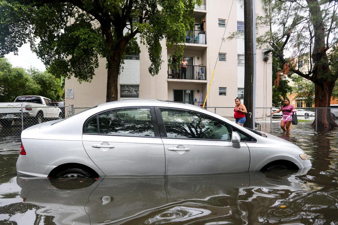 A couple of women walk by a car parked on the flooded corner of SW 8 Avenue and 4th Street as neighbors watch from a balcony in the neighborhood of Little Havana in Miami, Florida, on Saturday, June 4, 2022.