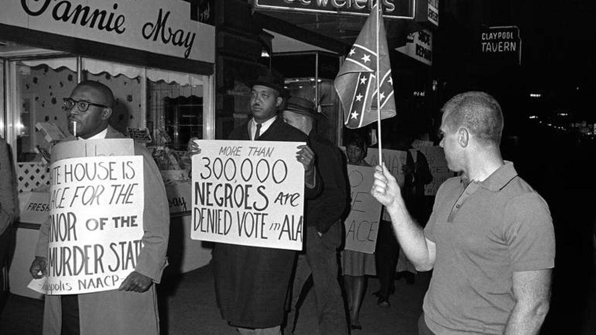 A man holds a Confederate flag as demonstrators, including one carrying a sign saying: “More than 300,000 Negroes are Denied Vote in Ala”, demonstrate in front of an Indianapolis hotel where then-Alabama Governor George Wallace was staying on Apr. 14, 1964.