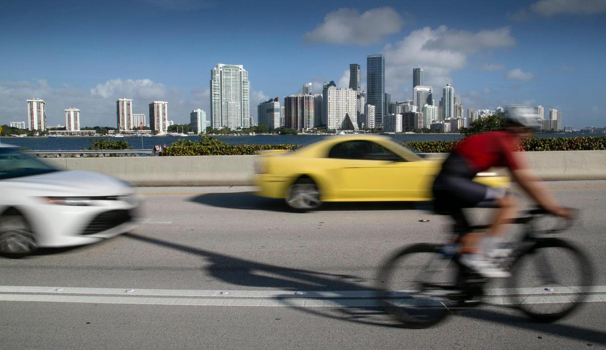 The downtown Miami skyline is visible in the background as vehicles pass a cyclist on the William Powell Bridge.