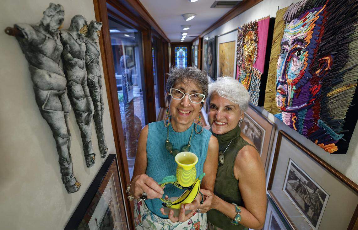 Lisa Palley (standing with a banana goblet by artist Robert Levin) and Donna Kass stand in the Palley family home’s “portrait gallery” in Miami on Sept. 10, 2025, amid artwork made with color pencils, shoelaces, ceramics, water colors, pen and ink, batik and metal screws.