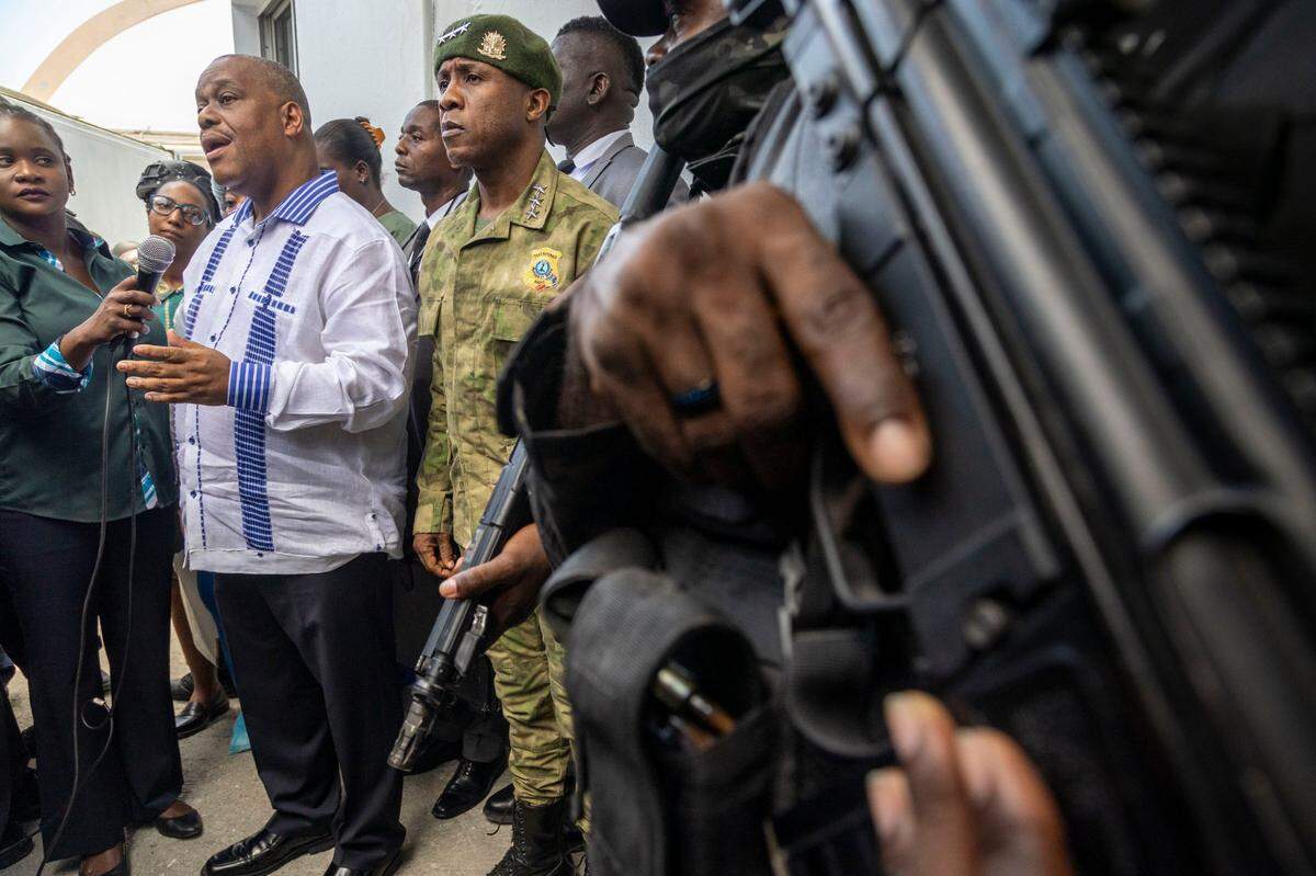 Port-au-Prince, Haiti, August 27, 2024 -A Police guard keeps his trigger finger at the ready as Haitian Prime Minister Gary Conille, left, speaks to the press along with Haiti Police Chief Rameau Normil, right, at Hospital Bernard Mevs where they went to visit Haitian police officers who have been wounded while battling gangs in Port-au-Prince.