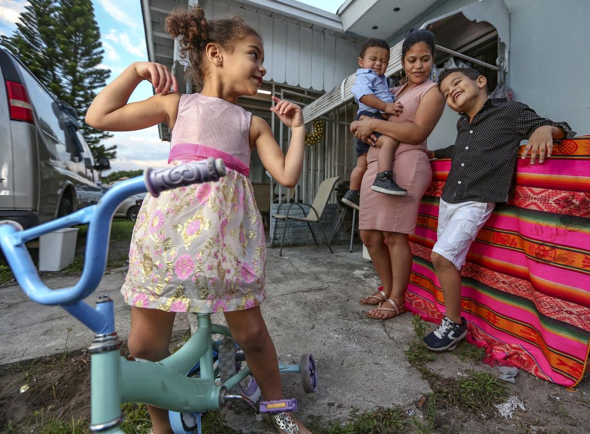 Greila Javier de Benavides holds her son Erick, 1, as her daughter Solimar, 4, at left and Alexis, 6, at right, look on while outside their home in Miami on Dec. 4, 2018. The boys are visually impaired and need specialized glasses. The family has financial difficulties. Meanwhile, mold issues in their home have forced them to get rid of a couch, two mattresses, clothing and several toys.