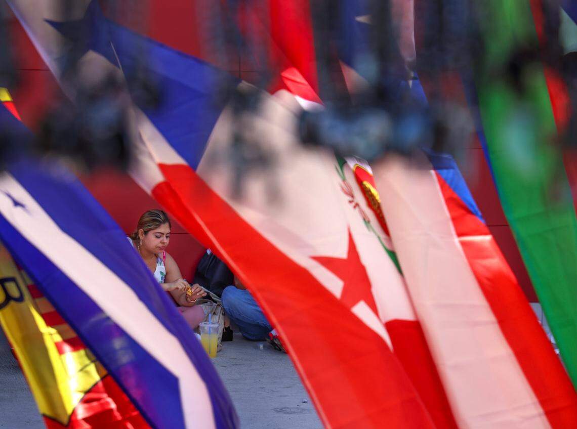 Among the music, merchandise, and food, a woman and her companions enjoy the street cuisine of Calle Ocho framed by flags and other items for sale on Sunday, March 12, 2023, at the Calle Ocho Music Festival.