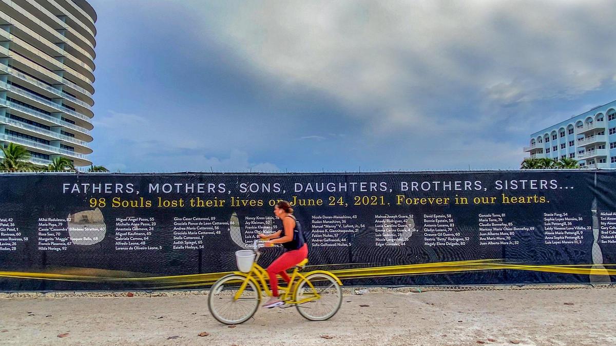 A woman bikes by a banner that honors the 98 victims of the Champlain Towers South condominium building collapse.
