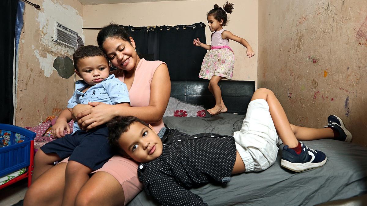 Greila Javier de Benavides, 26, holds her son Erick Benavides Javier, 1, and Abdiel Alexis Benavides Javier, 6, is seen at front right, as her daughter Solimar Benavides Javier, 4, jumps on the bed in the bedroom they all share at home in Miami on Dec. 4, 2018. Mold issues in their home have forced them to get rid of a couch, two mattresses, clothing and several toys.
