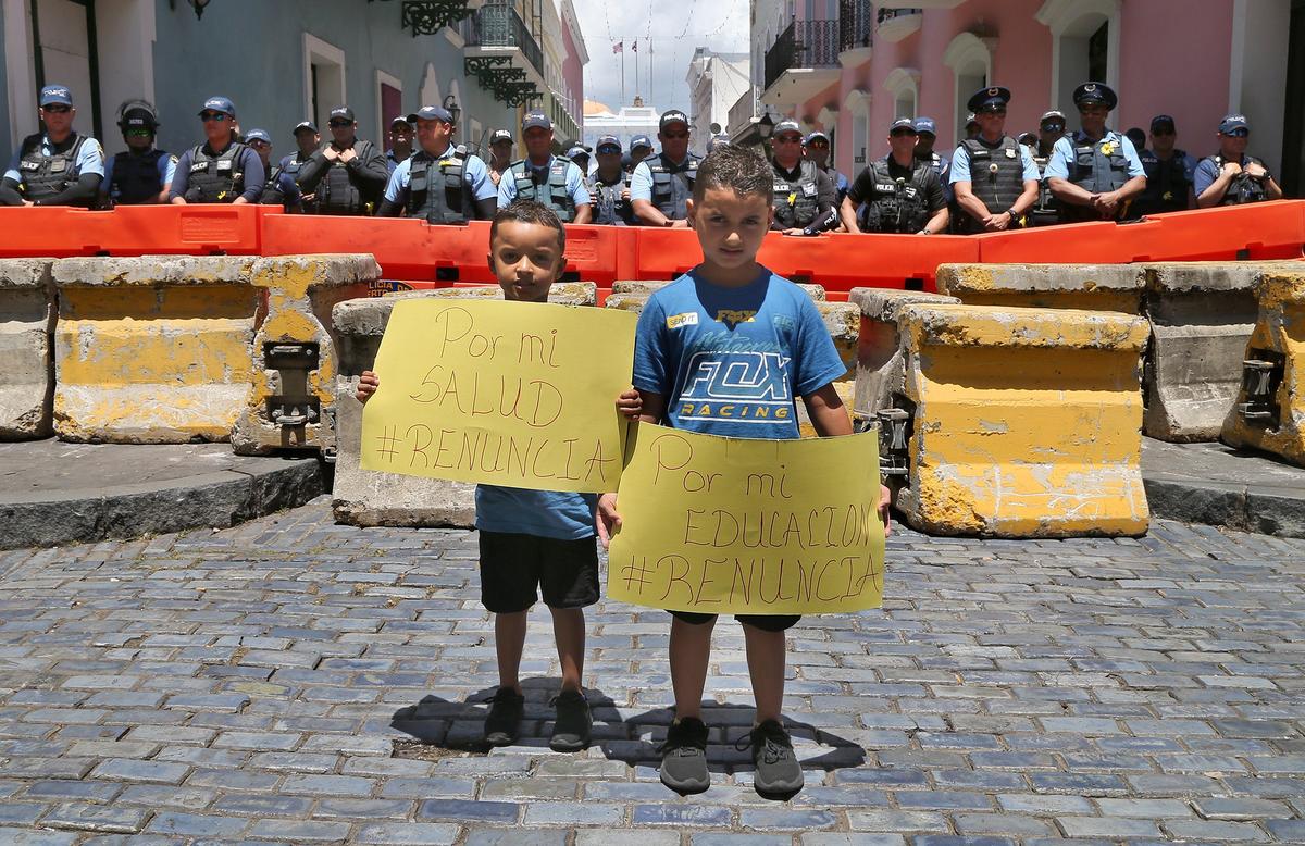 Kids Dylan, left, and Roy Salgado display signs Wednesday in front of a security perimeter set up by by local police in Old San Juan. The signs demand the resignation of Gov. Ricardo “Ricky” Rossello.