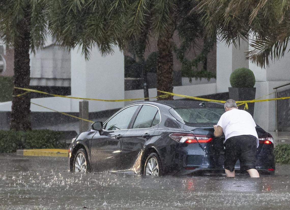 A person pushes a car through a flooded portion of Northeast 11th Street in downtown Miami as lightning and heavy rain falls over the area on Monday, June 19, 2023.