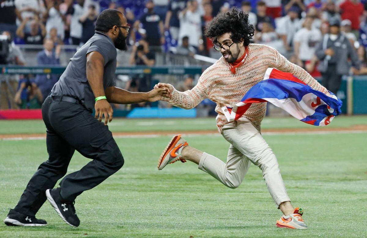 Cuban writer Carlos Manuel Alvarez carrying a Cuban flag with the words Patria y Vida runs on the field as Cuba infielder Andy Ibáñez (77) looks on during the game between the United States and Cuba at the World Baseball Classic semifinal at loanDepot Park in Miami, Fla. on Sunday, March 19, 2023.