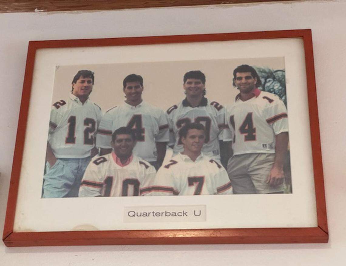 A wall of fame photo at The Big Cheese titled Quarterback U features University of Miami Hurricanes 1980s football stars #12 Jim Kelly, #4 Steve Walsh, #20 Bernie Kosar, #14 Vinny Testaverde, #10 George Mira Sr. and #7 Craig Erickson.