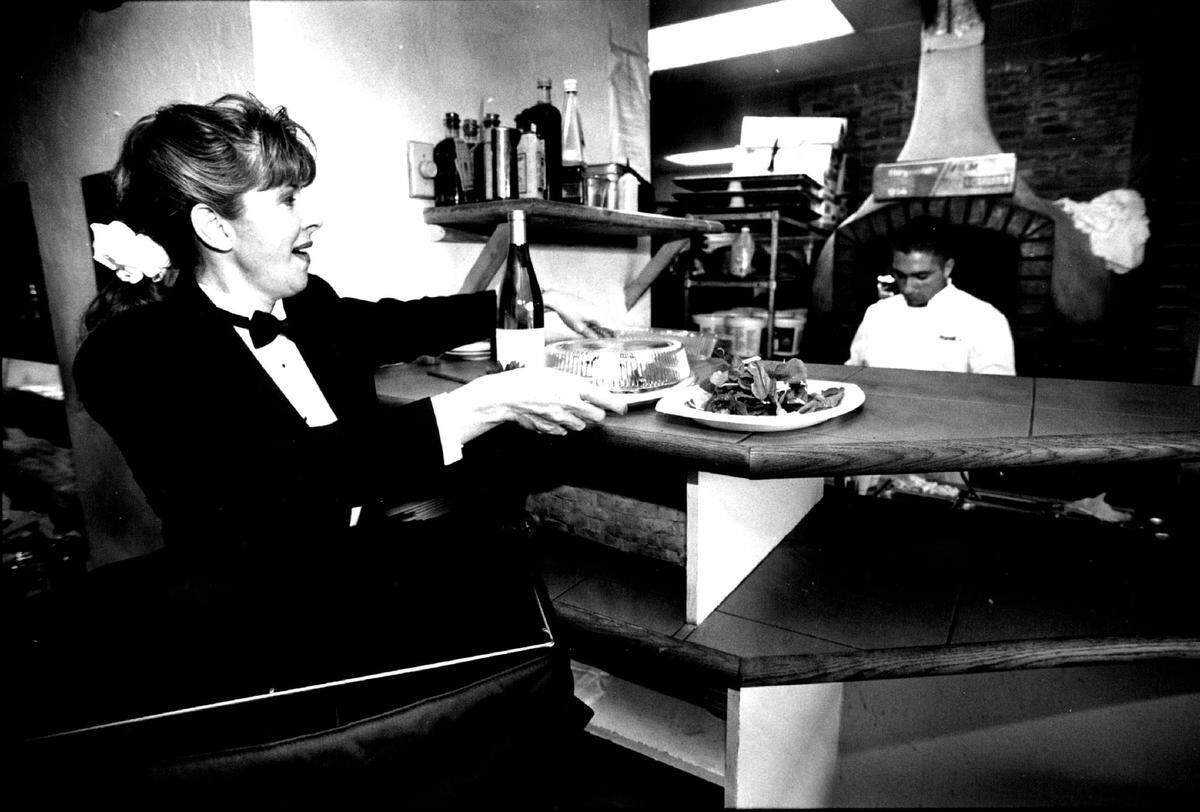 Tuxedo clad delivery serve Debi London picks up food prepared by line cook Mike Malki at Mark’s Place in North Miami.