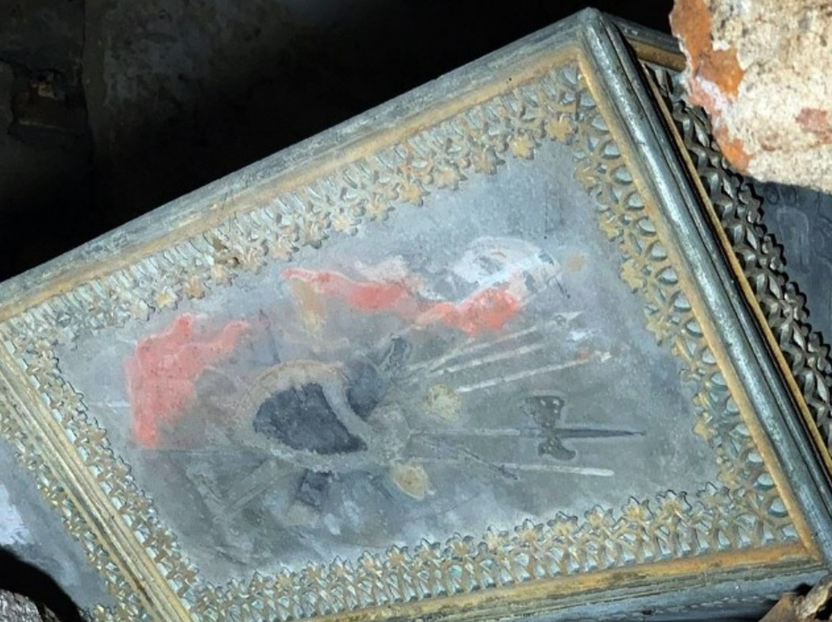 A detailed view of the coffin reveals a painting of a shield and weapons.