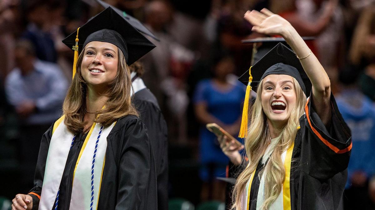 Gillian Drexler, right, greets her family in the stands as Charleigh Peters, left, leads the way down the aisle as they enter the Watsco Cente for UM’s spring commencement ceremonies Friday morning. Drexler majored in biochemistry and molecular biology and art, and minored in chemistry, and wants to become a dentist next.
