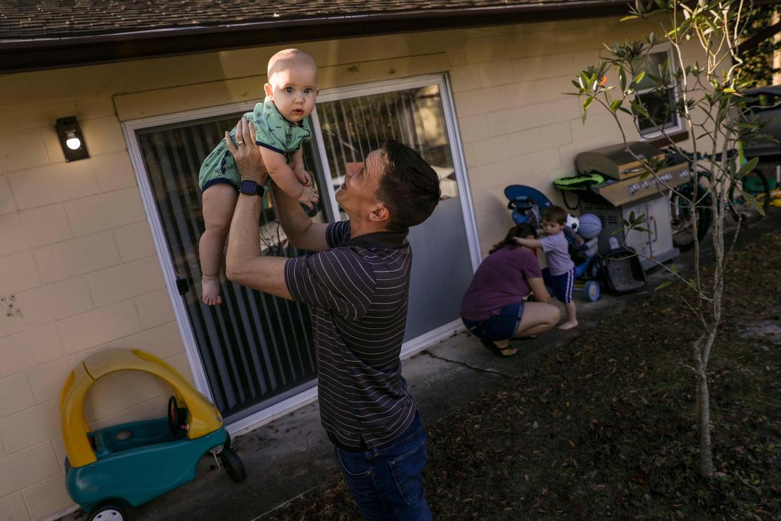 Jason Tallafuss, 42, carries Alexander, 9 months, while Danielle Tallafuss, 35, helps Benjamin, 2, puts his shoes on in the front yard of their family’s Oveido home on Monday, Feb. 21, 2022. After adopting Benjamin and before Alexander was born, the parents decided to terminate Danielle’s first pregnancy at 22 weeks upon learning that their son Nathaniel would have been born with an underdevelopment of the left side of the heart, to protect their child from the toll his condition would take on his life.