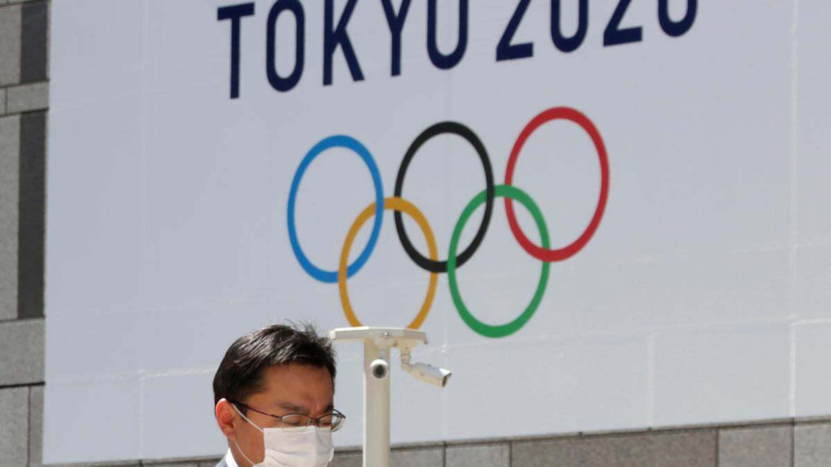 In this March 25, 2020, file photo, a masked man walks in front of a Tokyo Olympics logo at the Tokyo metropolitan government headquarters building in Tokyo. The Tokyo Olympics have been moved to 2021, but countless questions remain.
