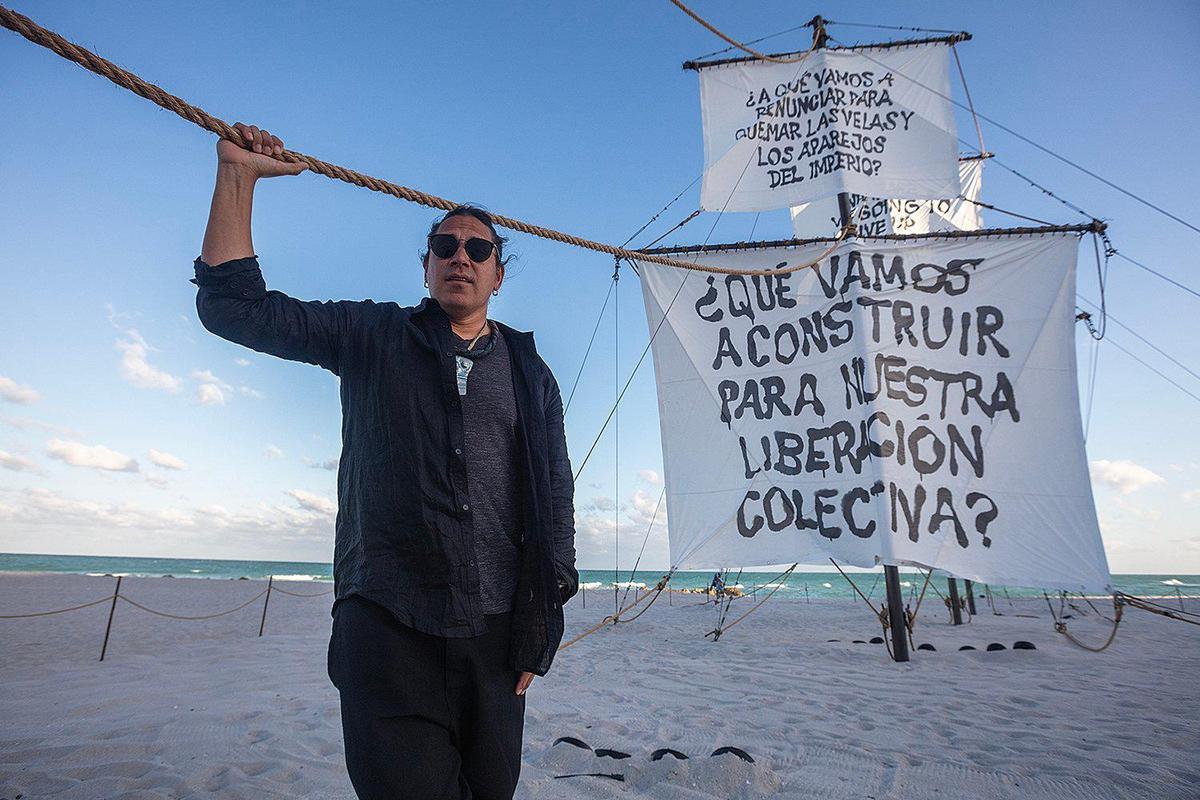 Artist Nicholas Galanin poses next to his art installation “Seletega,” a 90-foot-long sculpture of a wrecked Spanish galleon, partially buried in the sand, exhibited during Miami Art Week at the sands on Miami Beach.