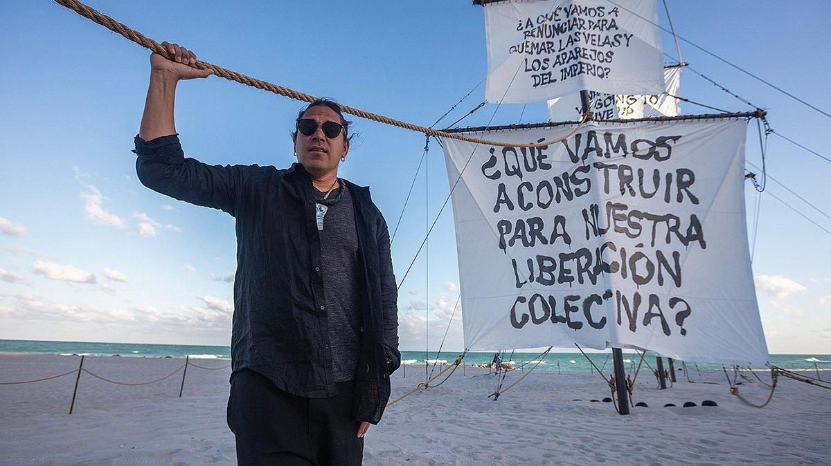 Artist Nicholas Galanin poses next to his art installation “Seletega,” a 90-foot-long sculpture of a wrecked Spanish galleon, partially buried in the sand, exhibited during Miami Art Week at the sands on Miami Beach.