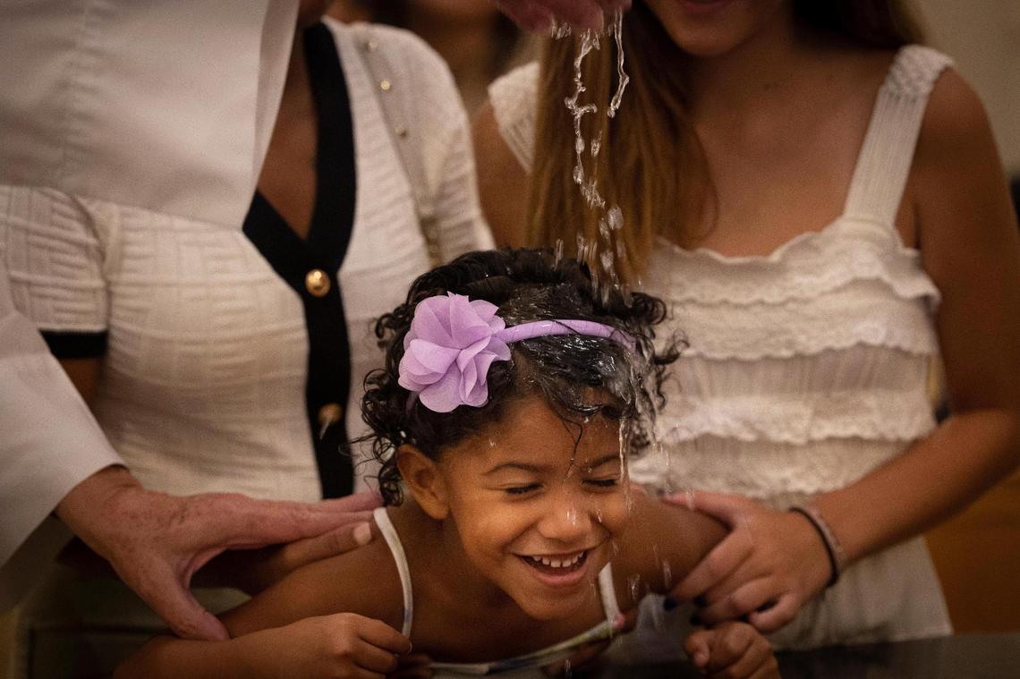 Karen Ugas’ daughter gets baptized by Father José Menéndez on Sunday, Sept. 10, 2023, at Corpus Christi Catholic Church in Miami. Many of the children baptized were migrants being welcomed into the church as part of an outreach effort by the Hermanos de la Calle, a organization that combats homelessness.