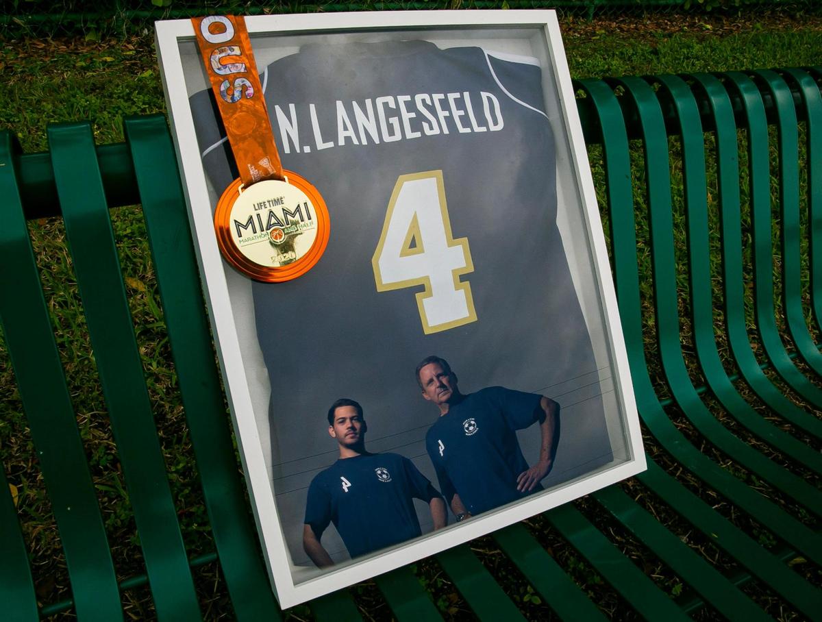 Martin Langesfeld and his father, Pablo Langesfeld, are reflected in a framed high school soccer jersey with a 2020 Miami Marathon medal belonging to Nicole Langesfeld in Doral on Wednesday, Jan. 26, 2022.