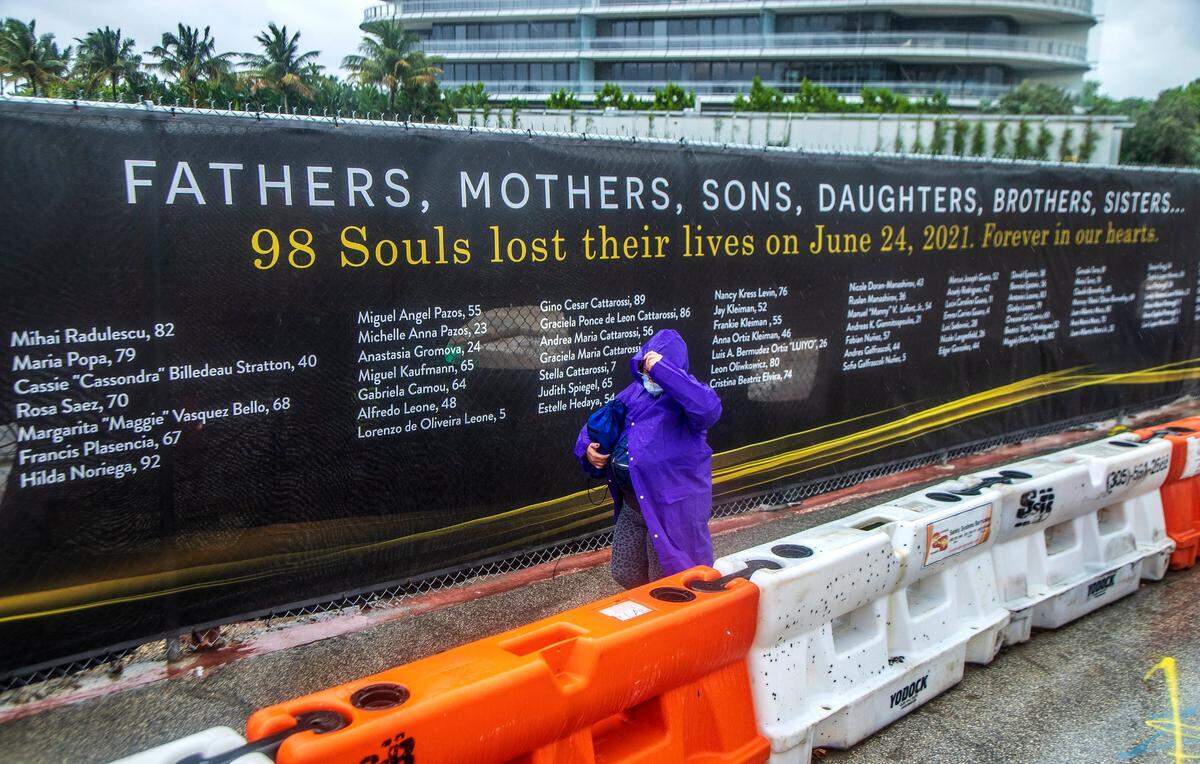 A woman takes cover under a raincoat as she walks by the memorial wall banner with the names of the 98 people that died at the Champlain Tower’s collapse at 8777 Collins Ave. in Miami Beach as the first slashes of rain from an incoming tropical disturbance reach Florida, on Friday June 3, 2022.