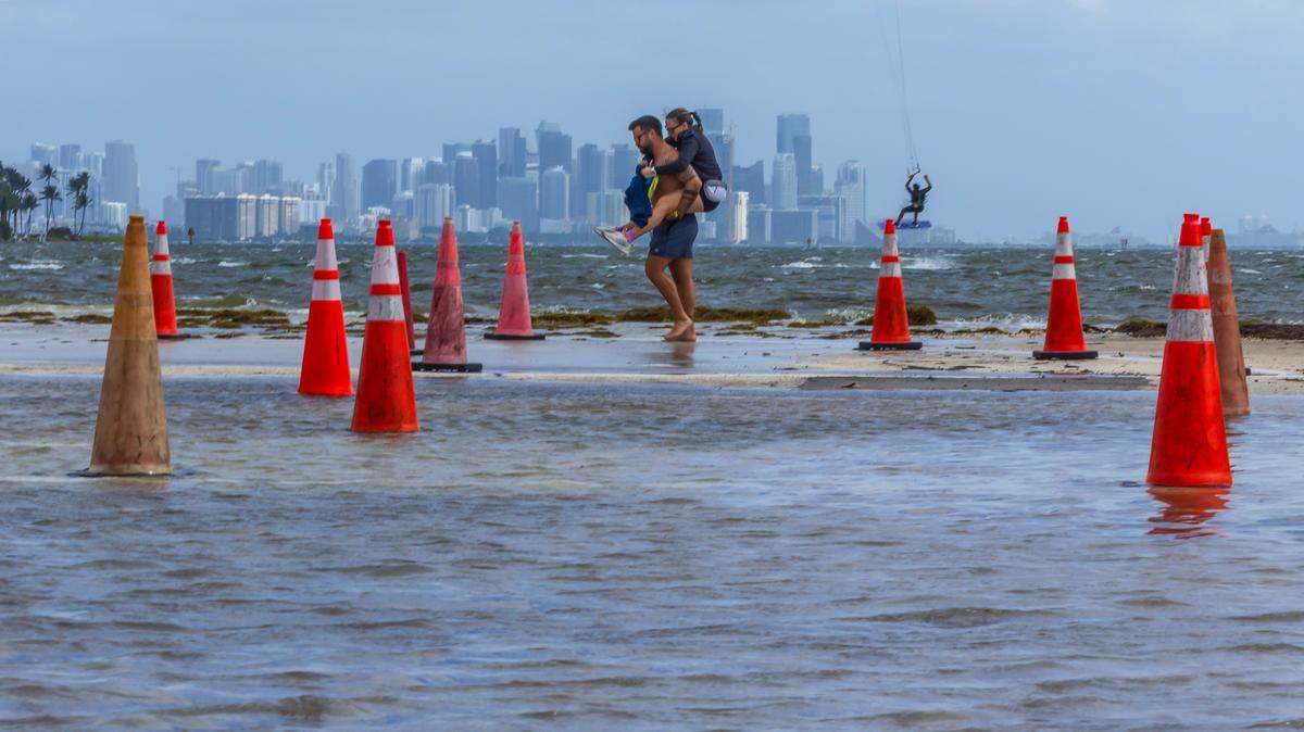 A Kite surfers take advantage of windy weather as a couple walk through saltwater from Biscayne Bay flooding the parking lot during the King tide peak at Matheson Hammock Park & Marina, in Miami, on Saturday October 19, 2024.