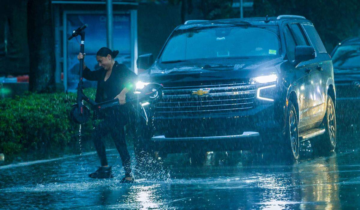 A woman carries her scooter in heavy rain as she crosses Biscayne Boulevard and 14th Street on Thursday, June 13, 2024.