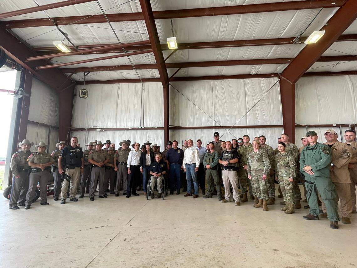 In this photo taken Saturday, July 17, 2021, in an airport hangar in Del Rio, Texas, Florida Attorney General Ashley Moody is near the center of the group to the left of Texas Gov. Greg Abbott while Gov. Ron DeSantis is on the other side of Abbott.