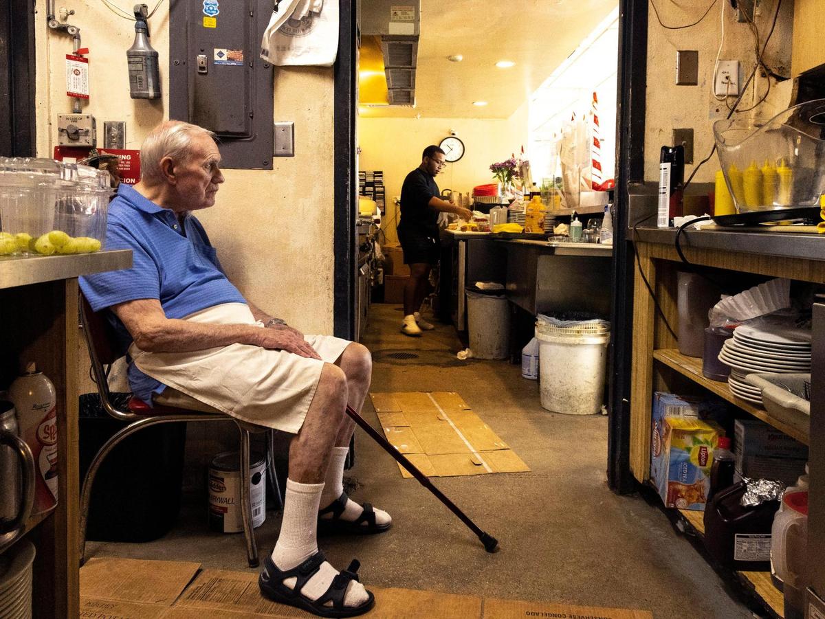 Bob Maguire, 85, sits as Corey Tennyson, 20, his partner’s son, works near the grill at Burger Bob’s on Monday, Dec. 20, 2021. It is a waiting game for Burger Bob’s owner, while he and his partner hope for a new lease in order to continue to operate the beloved Coral Gables dinner at its original location,