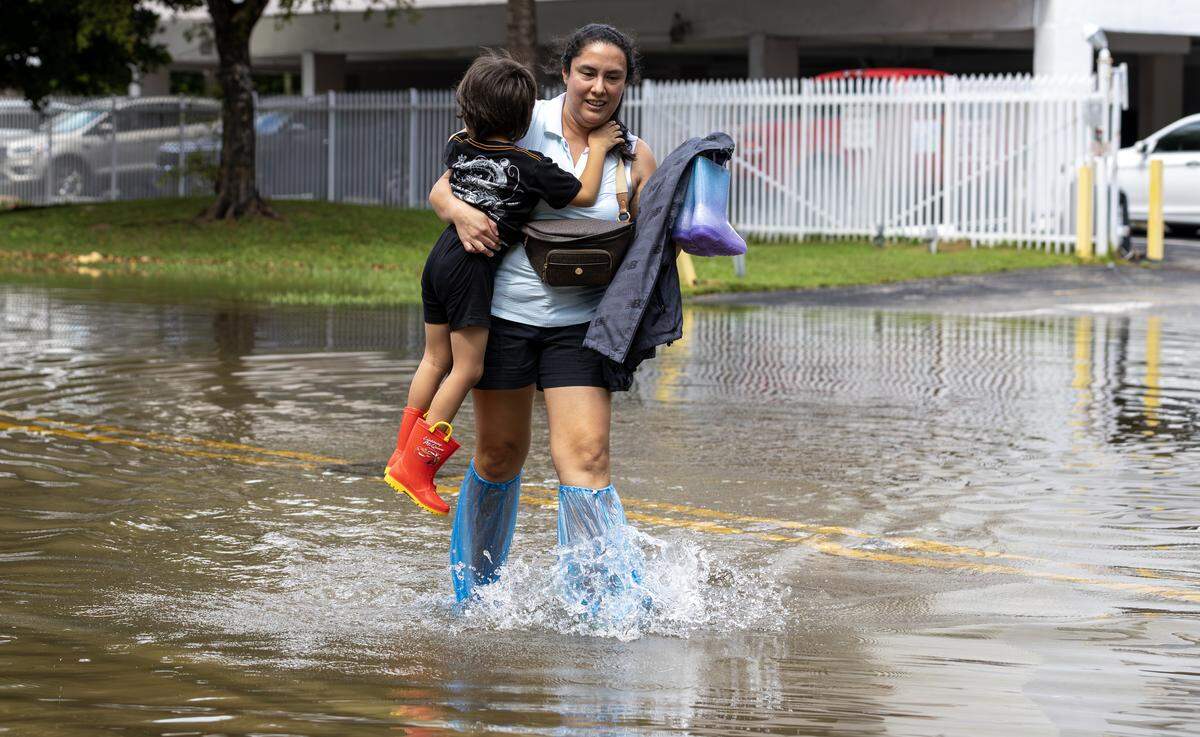 Madely Romero carries her 3-year-old son, Jared Santillana, through ankle-deep water as she crosses flooding caused by the king tide to reach a building on 250 180th Drive in Sunny Isles Beach, Fla., on Wednesday, Oct. 8, 2025.