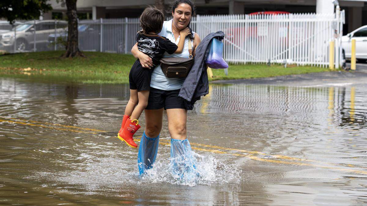Madely Romero carries her 3-year-old son, Jared Santillana, through ankle-deep water as she crosses flooding caused by the king tide to reach a building on 250 180th Drive in Sunny Isles Beach, Fla., on Wednesday, Oct. 8, 2025.