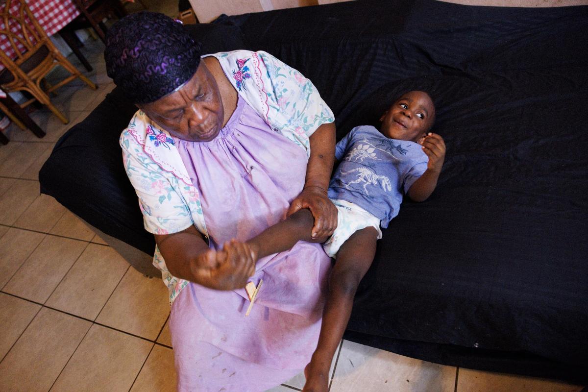 Mimose Martial, a 75 year-old grandmother who takes care of her grandchildren and adult daughter with down syndrome, checks on the foot of her grandson, Samson, 2, after he started crying. The grandchildren, all three of them being toddlers under 3, run around the home.