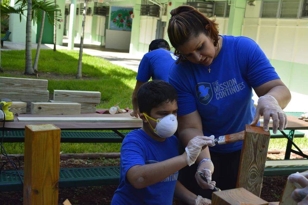 Derek Auguste’s wife, Kelly, and their son Donovan while working with The Mission Continues to stain an outside classroom desk.