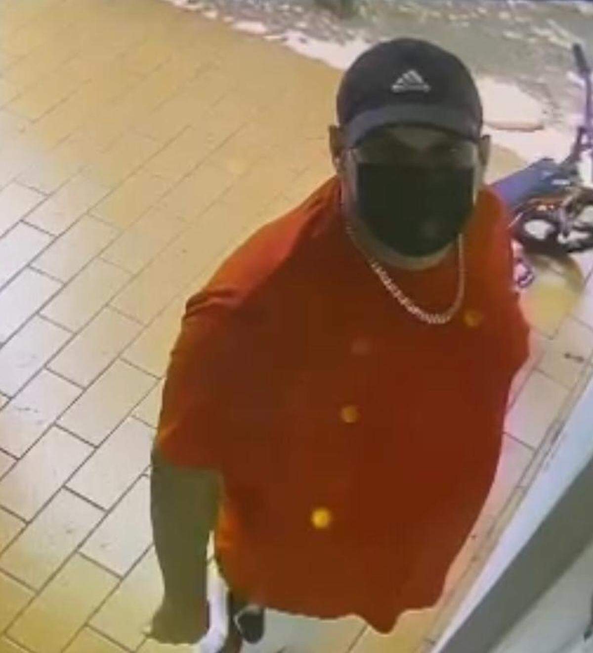 A man wearing a black hat, facial mask and red shirt looks at a security camera outside a home that Miami police say he and another man burglarized Monday, Jan. 23, 2023.