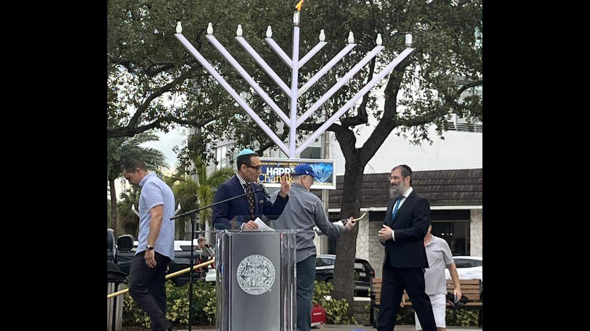 Coral Gables Mayor Vince Lago leaves the stage as Coral Gables attorney Spencer Aaronfeld, at the podium, greets Chabad of Coral Gables co-director Rabbi Avraham Stolik during the Menorah lighting celebration at Ponce Circle Park, Sunday, Dec. 14, 2025.