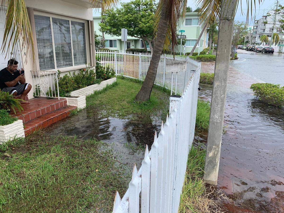 A Miami Beach man smokes a cigarette and video chats with a friend on his porch, the only dry spot on his street after a king tide-related flood. Sea rise related flooding is expected to become more common in the future, and flood-prone homes will either be redeveloped or relocated.