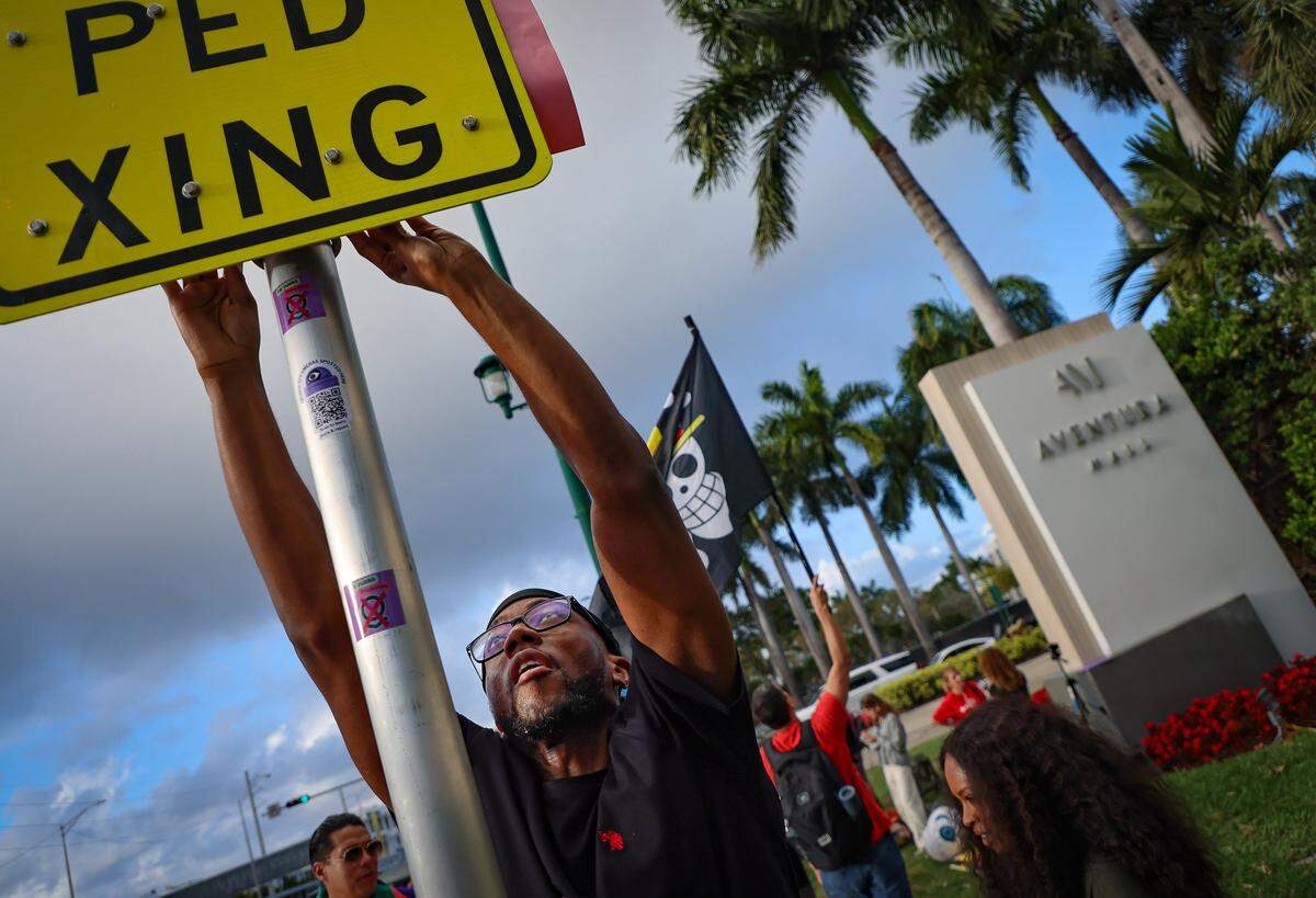 A protestor stretches to place sign on a pole as a small group gathered around the mall entrance, wielding bright signs that conveyed messages calling for increased accountability and transparency from the tech giant on Tuesday, March 3, 2026 at the entrance of Aventura Mall in Aventura, Florida.