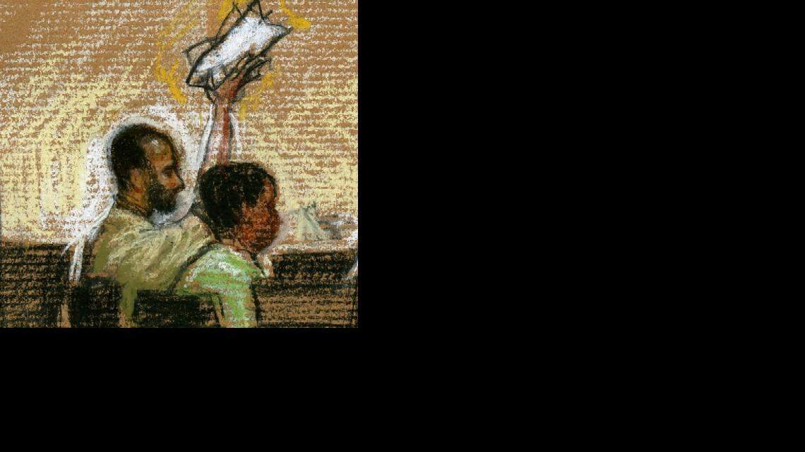 Accused al Qaeda propagandist Ali Hamza al Bahlul waves a boycott sign at his May  7, 2008 arraignment on war crimes charges at Camp Justice, the military commissions complex, at the U.S. Navy base in Guantánamo Bay, Cuba, as shown in a courtroom sketch submitted to U.S. military scrutiny before release. At his right is a Pentagon paid translator.