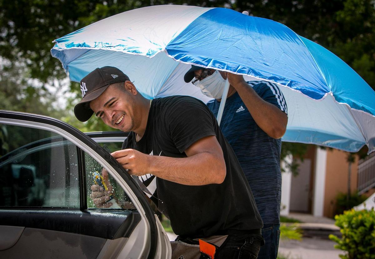 Miami Luxury Window Tinting employees Luis Rivas, left, installs window tinting on a car while his assistant, Juan Perez, right, holds a beach umbrella to protect both of them from the searing sun at Shoma Homes, corner of Northwest 70th Avenue and 177th Street, Palm Springs North in Miami on Monday, June 21, 2021. Normally, window tinting is done in a closed area where workers are protected from the elements but because of the pandemic, Rivas has been going to the customers, and that involves working outdoors exposed to the heat and humidity.