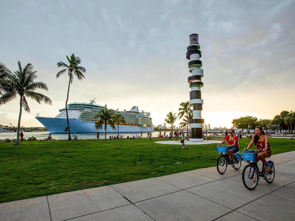 Two people bike down the sidewalk as Royal Caribbean International’s Freedom of the Seas cruise ship sets off down Government Cut past South Pointe Park for a simulated voyage leaving from PortMiami in Miami, Florida, on Sunday, June 20, 2021. In accordance with CDC requirements, the purpose of the simulation is to observe the cruise lines multilayered health and safety measures.