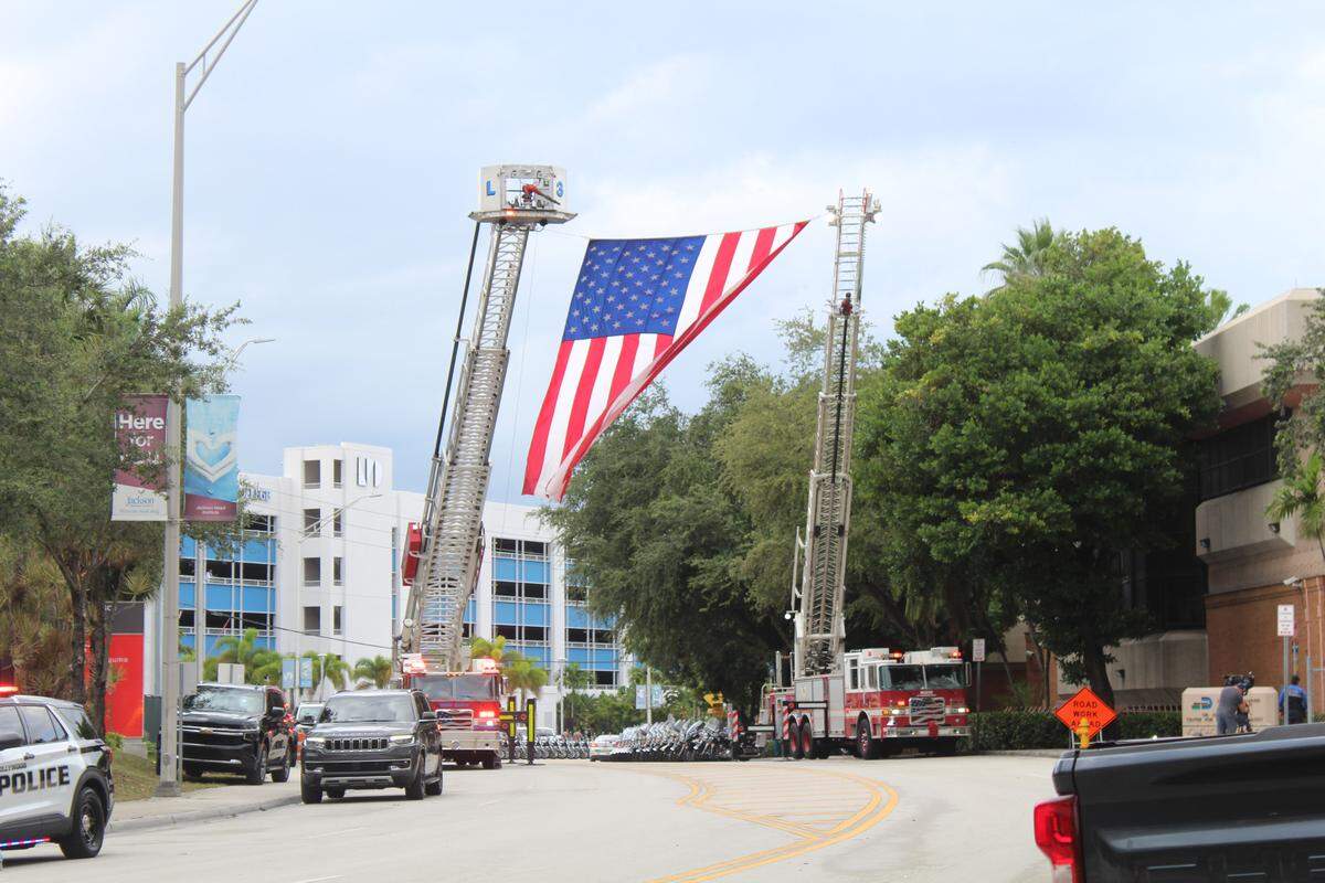 A large American flag hangs from the ladders of two Fire Rescue trucks parked at Jackson Memorial Hospital Tuesday, Oct. 28, 2025, in honor of Miami Beach police Officer David Cajuso, who was killed in a motorcycle crash earlier in the day on Interstate 75.
