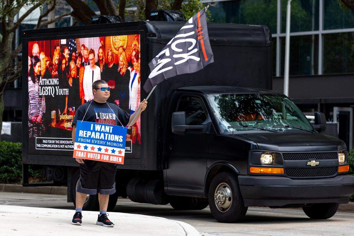 A protester from Indiana waves an anti-Biden flag and wears. a sign that reads, “I WANT REPARATIONS FROM EVERY IDIOT THAT VOTED FOR BIDEN” as a truck playing anti-DeSantis video passes by outside the Four Seasons Hotel in the Brickell business district of Miami, Florida, on Wednesday, May 24, 2023.