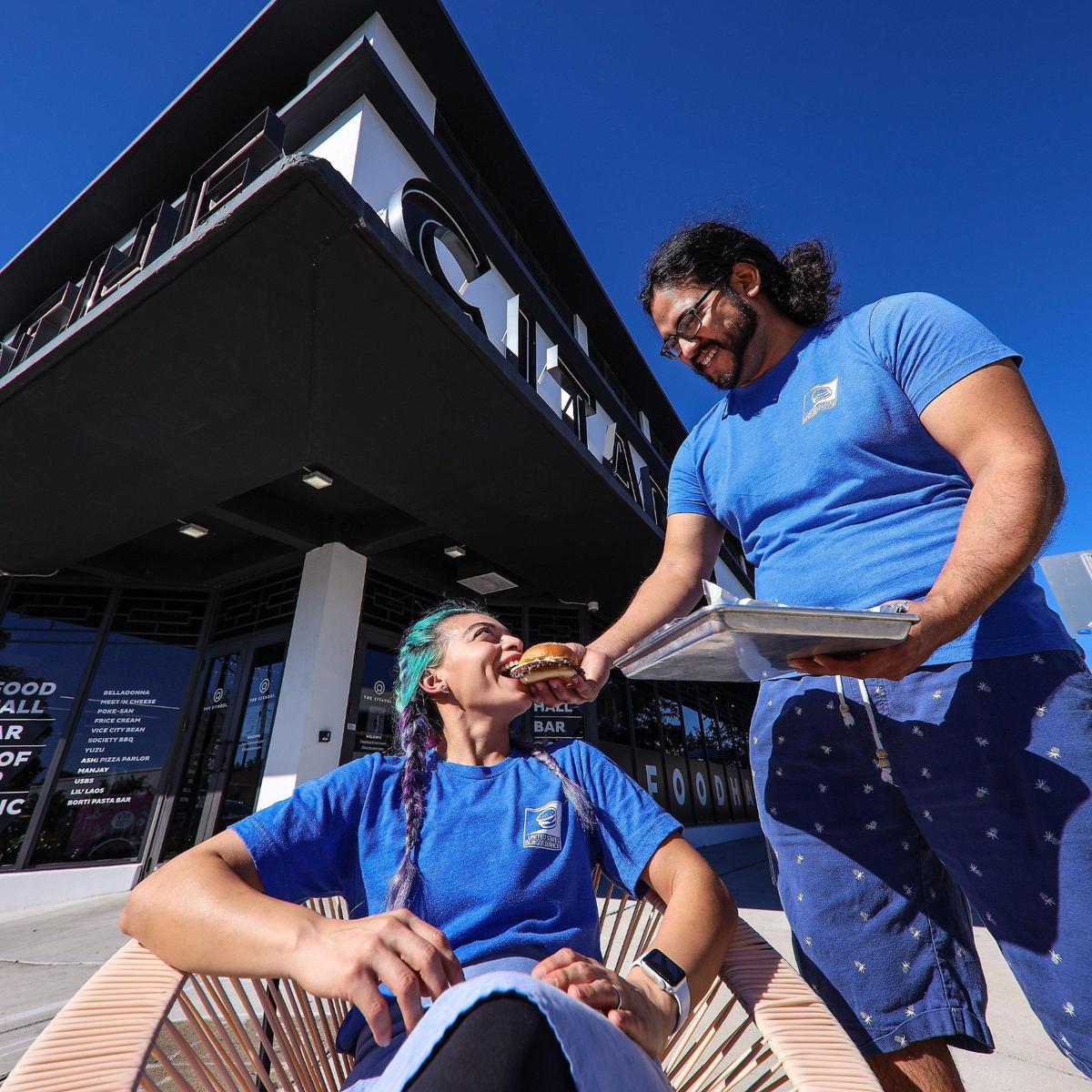 Keily Vasquez, left, takes a bite of a USBS (United States Burger Service) hamburger from her husband Mike Mayta, right, as they stand outside The Citadel food hall.