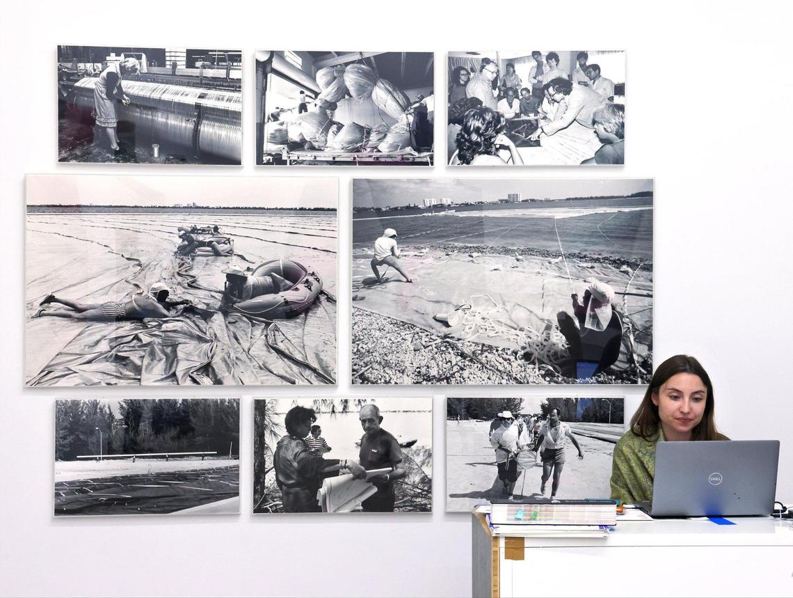 NSU Art Museum collection registrar Caroline McNabb sits at her desk next to black-and-white photographs documenting the process of creating “Surrounded Islands,” including the production of the pink fabric and the manpower needed to install it on the water.
