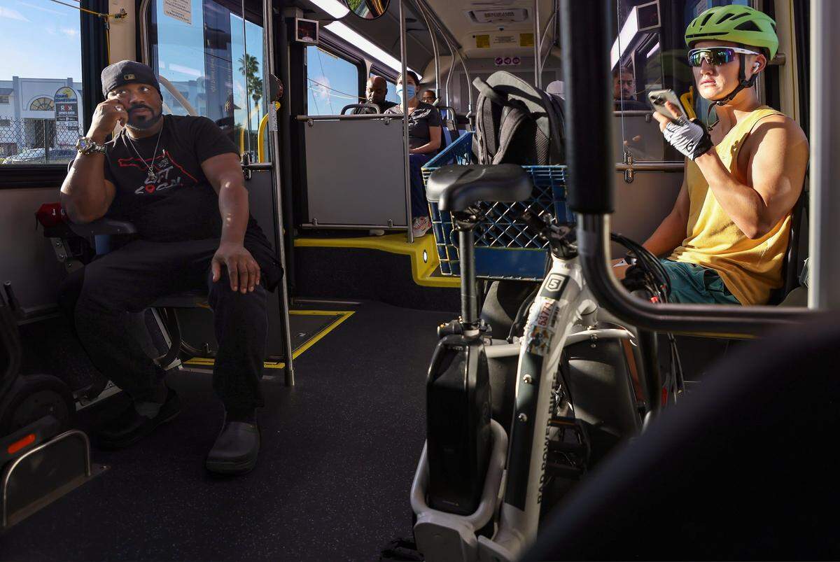 Commuters Vernon Huggins, 49, left, left, and Kevin Larson, 28, right, both use electric devices as part of their transit experiences on the first day of Miami-Dade's $300 million rapid-transit bus line known as “Metro Express” on Monday, October 27, 2025, just west of Richmond Heights on Coral Reef Drive and U.S. 1. The pair are disappointed in the travel time of the Metro Express buses as many first day riders complained about crowded buses and long travel times due to red lights on the express lane.